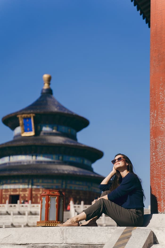 Una turista sentada en los escalones de mármol del Templo del Cielo en Pekín, disfrutando del sol con lentes oscuros, con el Salón de Oración por la Buena Cosecha difuminado al fondo bajo un cielo azul despejado.