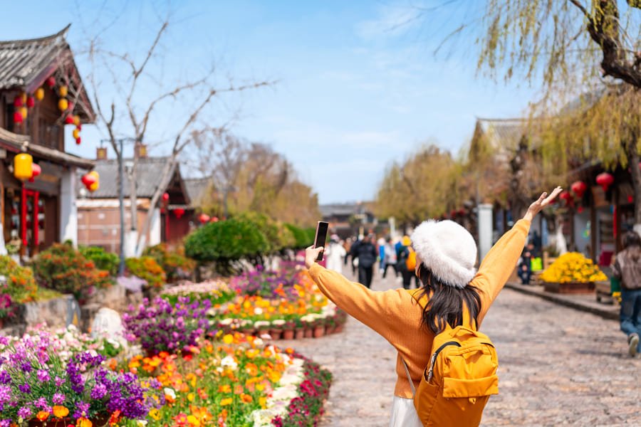 Vista desde atrás de una turista con suéter naranja, mochila amarilla y gorro blanco de felpa, que camina con los brazos abiertos por una calle peatonal empedrada en el casco antiguo de Lijiang, China. La calle está flanqueada por arquitectura tradicional de madera con faroles rojos y coloridos parterres de flores.
