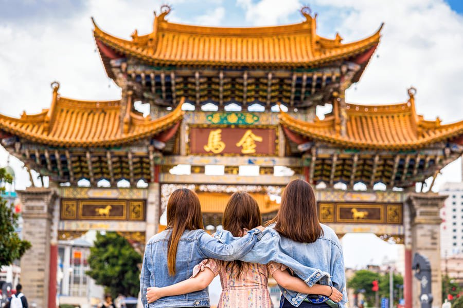 Vista desde atrás de tres mujeres jóvenes abrazadas por los hombros mientras contemplan una gran puerta o arco tradicional chino (Paifang) de color dorado y detalles ornamentados bajo un cielo parcialmente nublado.