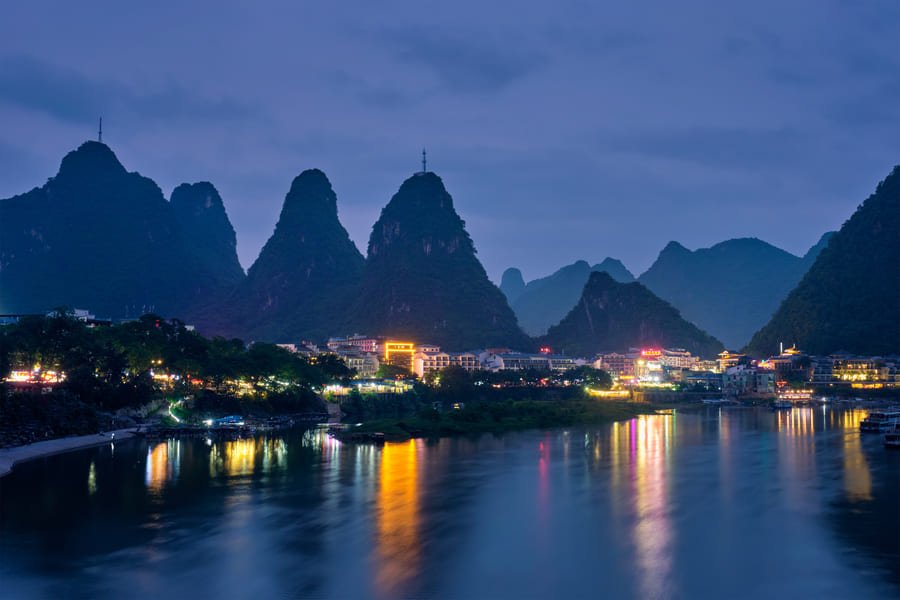 Vista panorámica nocturna de la ciudad de Yangshuo, en China, situada a lo largo del río Li. El primer plano está dominado por las tranquilas y profundas aguas del río, que actúan como un espejo para las vibrantes luces de la ciudad. El horizonte está jalonado por las icónicas montañas kársticas de piedra caliza, que se elevan imponentes en formas cónicas y se destacan contra el cielo azul-púrpura del crepúsculo.