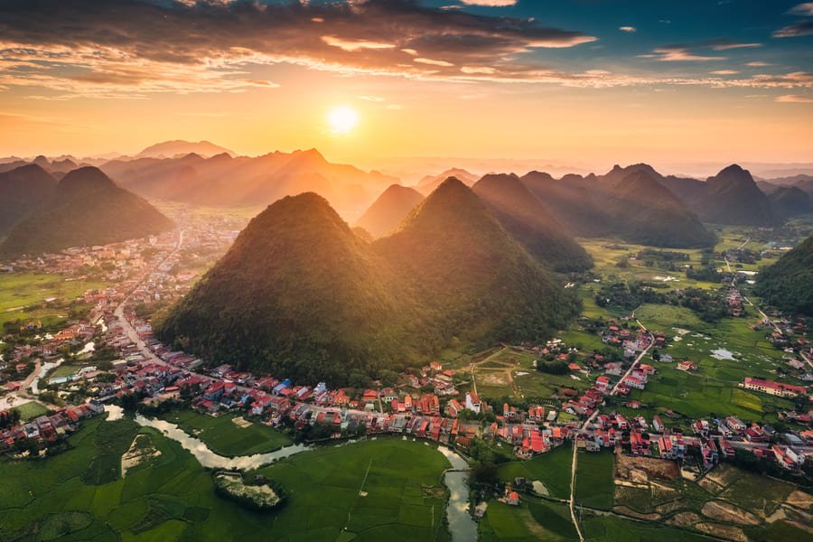 Vista aérea panorámica del paisaje kárstico de Yangshuo al amanecer. El sol brillante en el horizonte ilumina el cielo con tonos dorados y anaranjados, proyectando rayos de luz entre numerosas colinas cónicas y montículos verdes. A los pies de las montañas se extiende un valle con campos de cultivo verdes, un pequeño río serpenteante y un pueblo pintoresco con casas de tejados rojizos distribuidas a lo largo de los caminos.